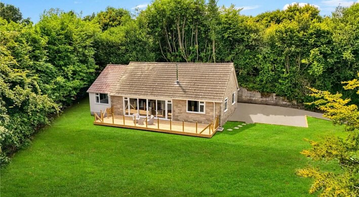 Aerial view of a small, single-story house with a sloped roof, surrounded by lush green trees and grass. The house has a deck with outdoor seating and large windows, set in a spacious yard under a clear blue sky.